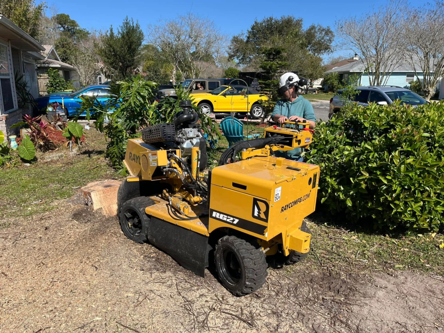 AGES Tree Service operator running Rayco RG27 stump grinder next to bushes in residential neighborhood