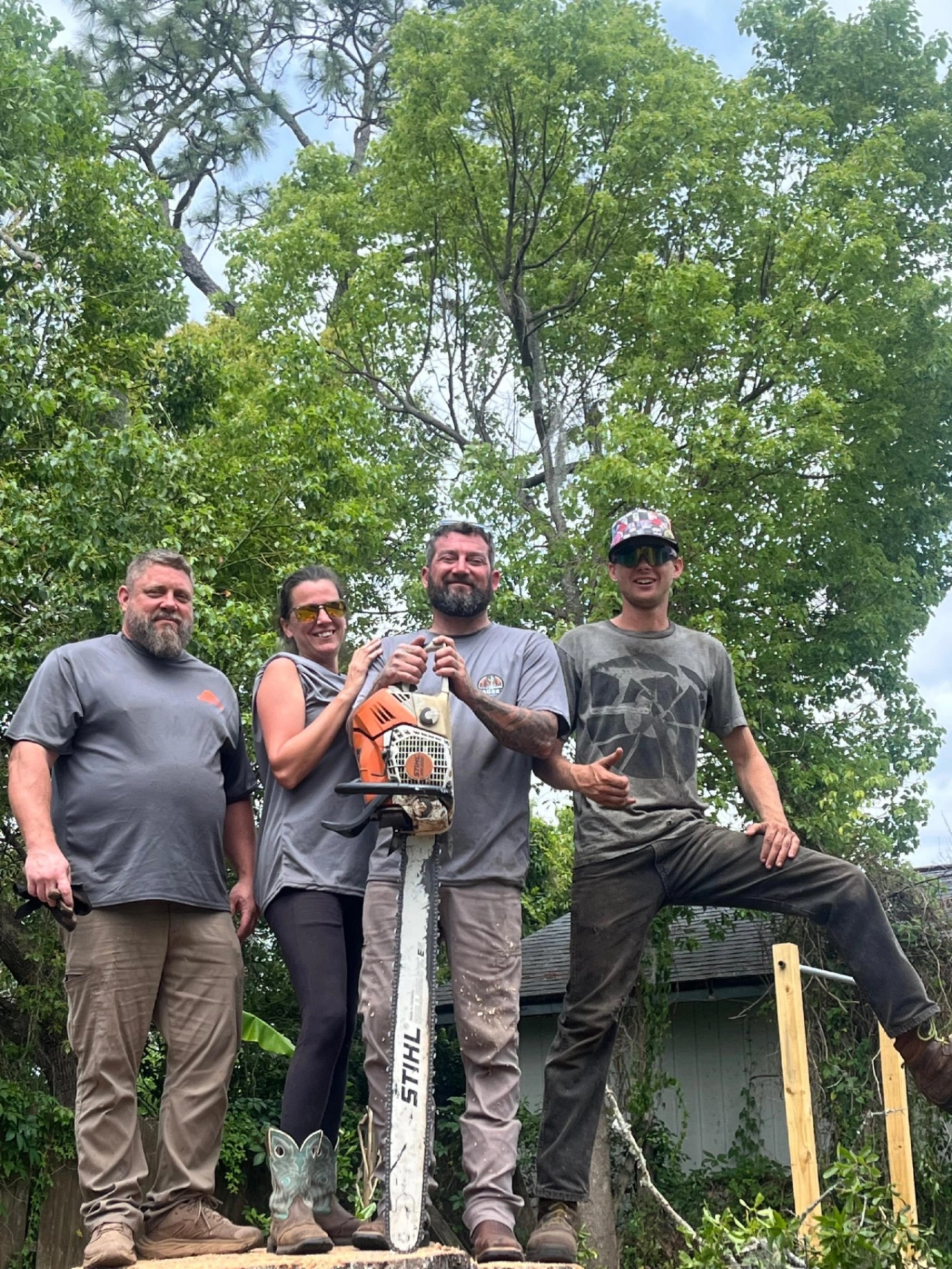 AGES Tree Service crew of four standing on a fresh-cut stump holding a STIHL chainsaw in Orange Park FL
